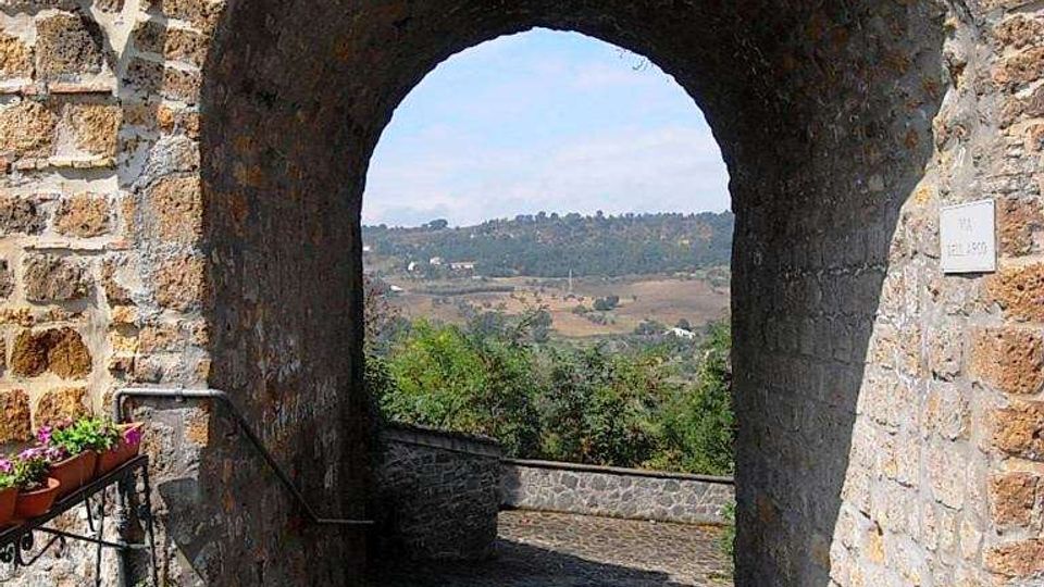 Arco in pietra di una porta medievale con pavimentazione in selciato e mura laterali, che incornicia la vista su una valle con campi e colline in lontananza. Sul lato sinistro si vedono una fioriera e un corrimano lungo la rampa in discesa.