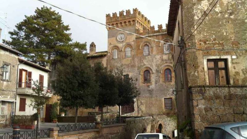 Veduta di un borgo con strada stretta e case in pietra, con auto parcheggiate ai lati. Sullo sfondo si distingue un edificio storico con facciata in mattoni e pietra, finestre ad arco, torre merlata e un piccolo orologio, circondato da alberi e cavi elettrici sospesi.