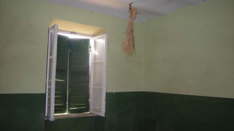 Interno di una stanza spoglia con pareti bicolori verde chiaro e verde scuro e soffitto bianco. Una finestra con persiane verdi e ante bianche aperte lascia entrare luce, con ganci e un fascio di paglia appeso al soffitto.