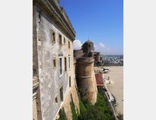 Veduta laterale di una fortezza costiera in muratura, con torre cilindrica e mura in pietra e intonaco, affacciata su un’area aperta vicino al mare. Sullo sfondo si vede un porto turistico con numerose barche e il litorale sotto un cielo sereno.