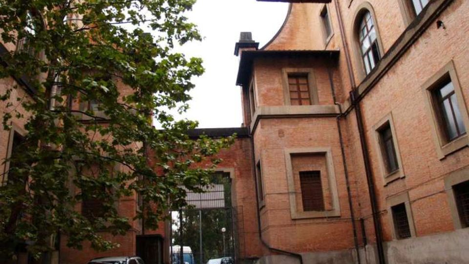 Cortile interno tra edifici in mattoni a più piani, con finestre ad arco e grondaie, affiancato da un grande albero. Un vialetto conduce a un cancello metallico verso la strada, con alcune auto parcheggiate ai lati.