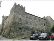 Edificio storico in pietra simile a un castello, con torre angolare merlata, mura inclinate e finestre con persiane. Davanti si vede un piccolo parcheggio con auto e una strada in pavé ai piedi della struttura.
