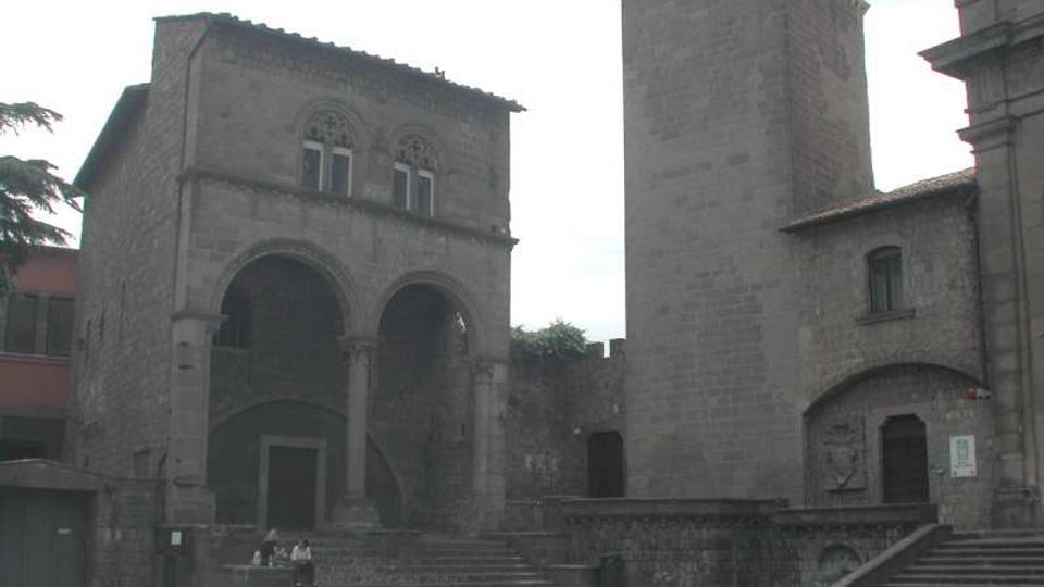 Piazza lastricata con complesso in pietra medievale, con un edificio a due arcate e scalinate d’accesso. Accanto si innalza una torre quadrata in muratura e sono visibili muri di cinta e facciate con piccole finestre.