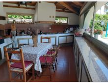 Cucina abitabile con soffitto in legno a vista, lunga penisola con piano chiaro e pareti piastrellate, affiancata da un tavolo con sedie in legno. Ampie finestre e una grande vetrata laterale illuminano l’ambiente e si affacciano sul giardino esterno.