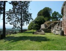 Prato alberato in un’area panoramica con panchine lungo una ringhiera, affiancato da resti di mura e strutture in pietra e mattoni. Sullo sfondo si vedono colline e un cielo sereno.