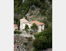 Complesso di edificio in muratura con piccola chiesa con campanile e tetti in coppi, situato su un pendio roccioso. Intorno si vedono alberi e vegetazione fitta, con pareti di roccia e una piccola terrazza murata davanti agli edifici.
