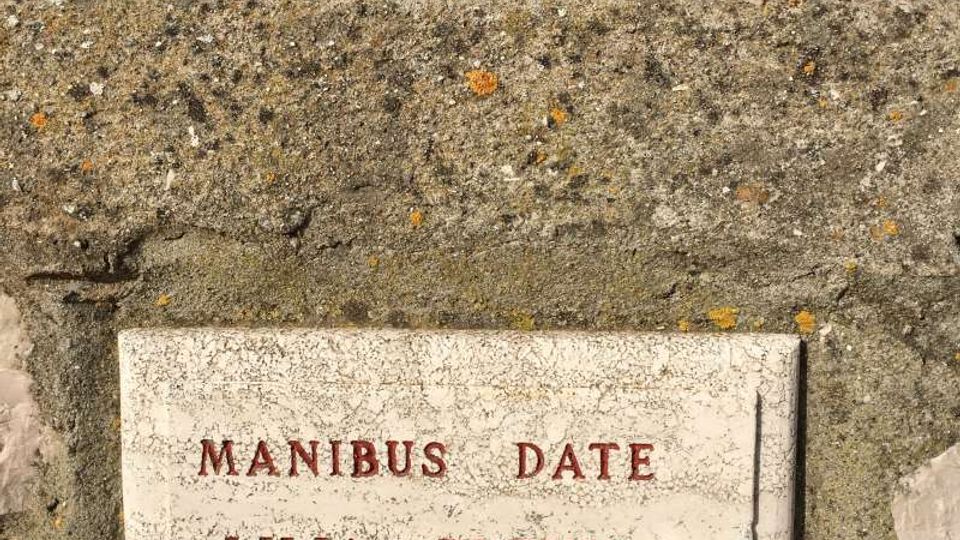 Lapide rettangolare in marmo incassata in un muretto di pietra, con iscrizione in rosso “MANIBUS DATE LILIA PLENIS VIRGILIO”. In alto si vedono erba e terreno, con cipressi sullo sfondo sotto un cielo azzurro.