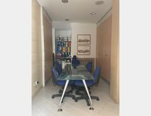 Piccola sala riunioni in ufficio con tavolo rettangolare in vetro e sedie girevoli blu, circondata da armadi in legno. In fondo si vede un’area con scaffali pieni di raccoglitori colorati, due stampe incorniciate sulla parete e faretti incassati nel soffitto.