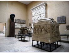 Sala museale con pareti beige e pavimento in pietra, con grandi reperti archeologici esposti su supporti neri, tra cui un sarcofago scolpito in primo piano. Alle pareti sono presenti rilievi e lastre decorative, oltre a un grande busto scuro su piedistallo.