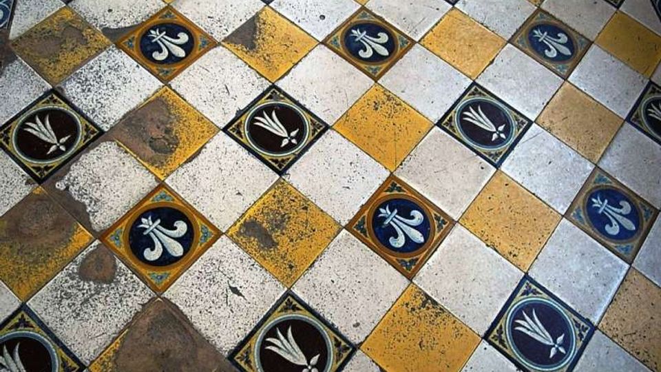 Pavimento in piastrelle a scacchiera con quadrati gialli e bianchi, intervallati da inserti decorativi blu e neri con motivi floreali. Le piastrelle mostrano segni di usura e macchie che evidenziano l’età della superficie.