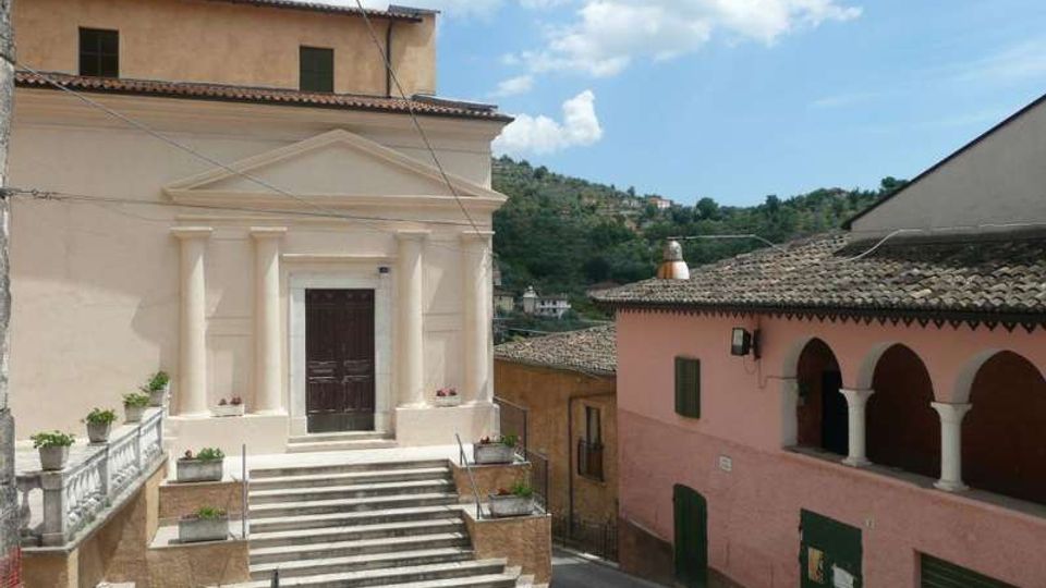 Piccola piazza di un borgo con una chiesa dalla facciata chiara, portale centrale e scalinata in pietra con ringhiere e fioriere. A destra si vede un edificio rosa con loggiato ad archi e tetto in coppi, sullo sfondo colline verdi sotto un cielo parzialmente nuvoloso.