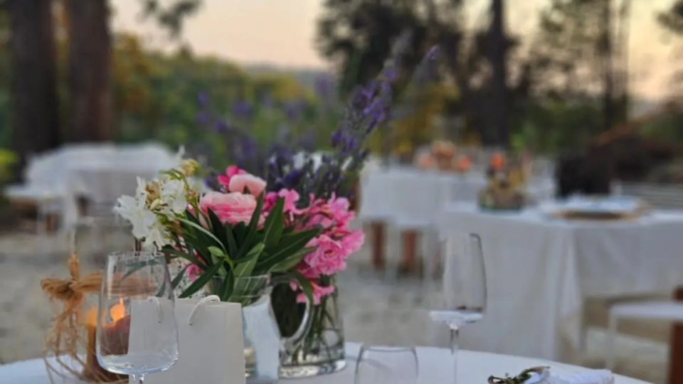 Tavolo apparecchiato all’aperto con tovaglia bianca, piatti, bicchieri e posate su sottopiatti intrecciati, decorato con un vaso di fiori e una lanterna. Sullo sfondo si vedono altri tavoli disposti in un’area verde con alberi al tramonto.