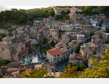Veduta dall’alto di un borgo collinare con case in pietra addossate tra loro e tetti in tegole, attraversato da strade strette che convergono in un piccolo incrocio. Intorno si vedono alberi e vegetazione sui pendii, con alcune abitazioni più recenti sullo sfondo.