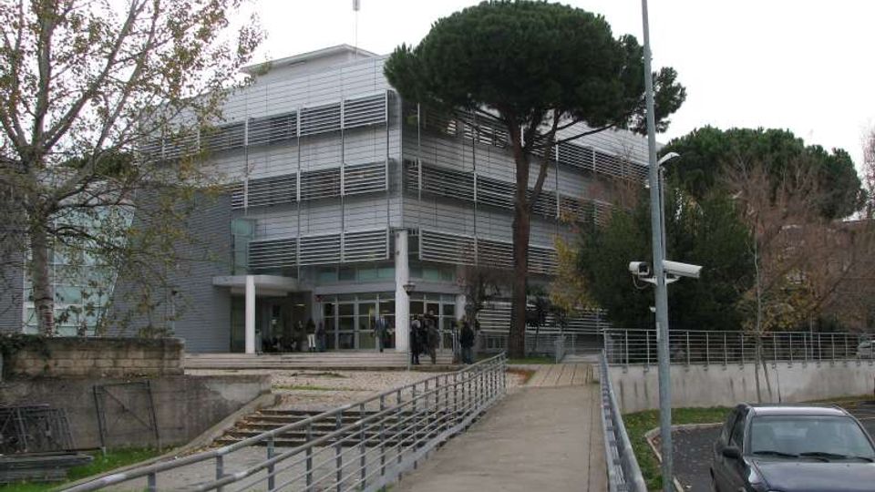 Edificio moderno a più piani con facciata in pannelli metallici e frangisole orizzontali, con ingresso vetrato raggiungibile tramite una rampa pedonale con corrimano. L’area esterna comprende alberi alti, un lampione e un parcheggio laterale.