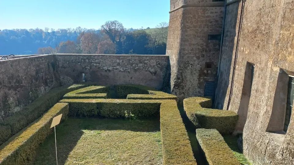 Giardino formale con siepi potate a labirinto all’interno di mura in pietra, accanto a un edificio storico con torre e facciata in muratura. Sullo sfondo si vedono colline alberate sotto un cielo sereno.