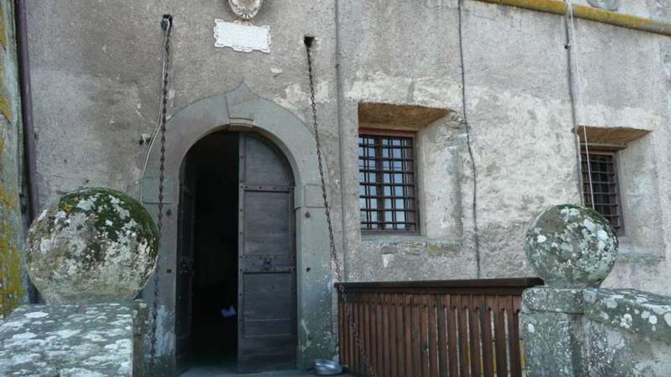 Facciata in pietra di un edificio storico con portale ad arco e grande porta in legno, affiancata da finestre con inferriate. Un breve ponte o passerella in legno con balaustra e pilastri in muratura sormontati da sfere conduce all’ingresso.