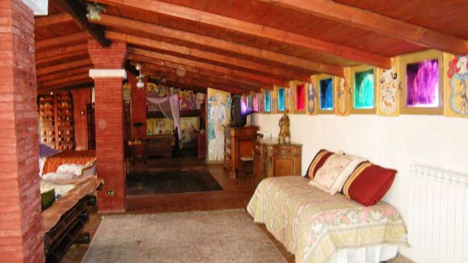 Mansarda arredata con soffitto spiovente in legno a vista e pilastri in mattoni, con pavimento in piastrelle e tappeti. Lungo una parete ci sono letti e mobili in legno, mentre l’altra è decorata da una fila di quadri colorati sopra un termosifone.