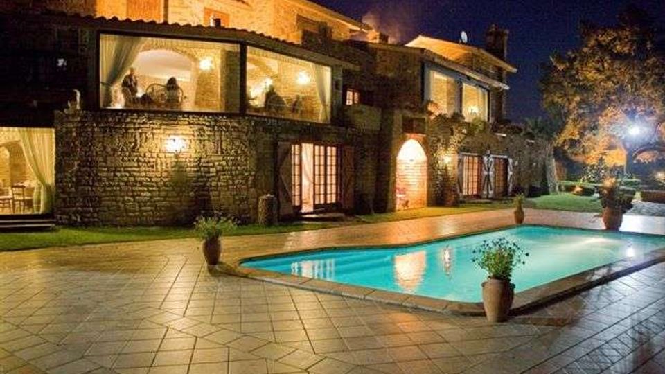 Villa in pietra illuminata di notte con ampie vetrate e porticati, affacciata su una piscina rettangolare con acqua turchese. Intorno alla piscina c’è una grande pavimentazione in lastre e vasi con piante, con giardino e alberi sullo sfondo.