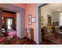 Interno di abitazione con corridoio dai muri rosa e pavimento in parquet, incorniciato da ampie porte con cornici grigie e tappeti persiani. A destra si apre un soggiorno con divani, poltrona, tavolino, libreria e lampada, illuminato da una finestra con tende chiare.
