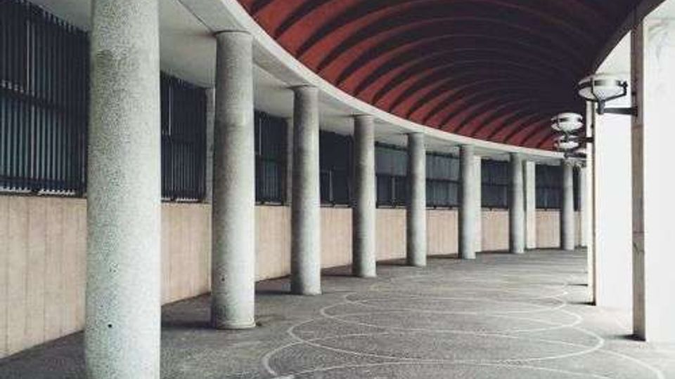 Porticato curvilineo con file di colonne cilindriche in cemento e soffitto a volta rivestito in pannelli rossi. Pavimentazione in pietra con motivi circolari incisi e lampade a parete lungo il lato esterno.