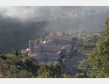 Vista dall’alto di un grande complesso monastico in pietra con campanile e tetti in tegole, disposto attorno a un chiostro interno. L’edificio è circondato da boschi e campi su un pendio collinare, con foschia nelle colline sullo sfondo.