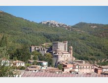 Veduta di un borgo collinare con un grande castello in pietra e una chiesa con campanile, circondati da case dai tetti chiari. Sullo sfondo si estendono pendii boscosi e creste montuose con un altro centro abitato in alto.