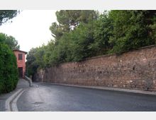 Strada curva in pavé accanto a un lungo muro in pietra sormontato da vegetazione fitta e alberi. Sul lato sinistro si vede un marciapiede che conduce a una piccola casa rosa con ingresso su una stradina laterale.