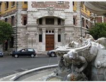 Fontana in pietra con una figura scolpita che versa acqua in una vasca semicircolare, in primo piano lungo una strada urbana. Sullo sfondo si vede la facciata di un edificio storico in muratura e mattoni, con ingresso ad arco e portone in legno.