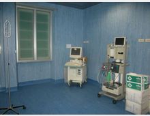 Stanza clinica con pareti e pavimento blu, finestra a due ante e attrezzature mediche su carrelli, tra cui un monitor e una macchina per anestesia o ventilazione con tubi e bombola. A lato si vedono un supporto per flebo e scatole di forniture impilate.