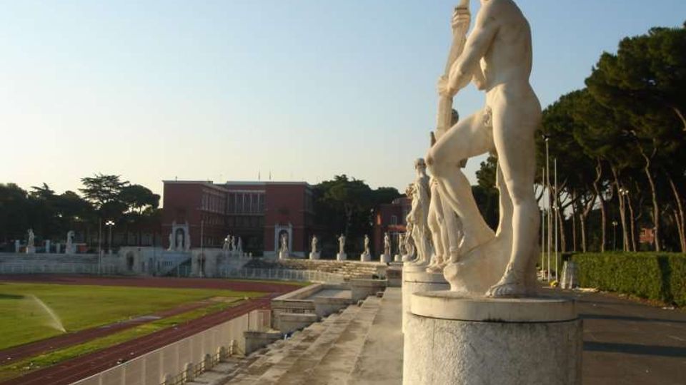 Viale di uno stadio all’aperto con gradinate in pietra e pista di atletica, fiancheggiato da una fila di statue classiche in marmo su basamenti. Sullo sfondo si vedono un edificio in mattoni rossi e alberi alti lungo il perimetro.