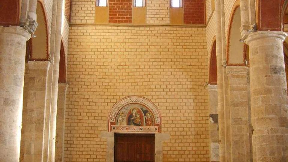 Interno di una chiesa in stile romanico, con navata centrale fiancheggiata da colonne in pietra e archi a tutto sesto. In fondo si vede una grande porta in legno con lunetta affrescata sopra, pareti in laterizio e piccole finestre alte, e pavimento a mosaico geometrico con file di sedie trasparenti ai lati.