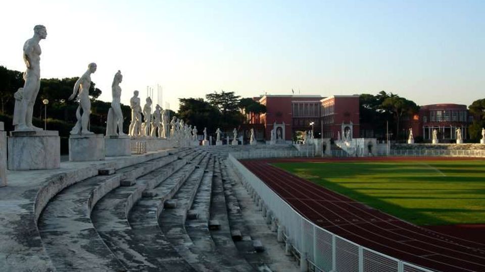 Gradinate in pietra di uno stadio all’aperto con una fila di statue classiche su piedistalli lungo il lato sinistro. A destra si vedono la pista di atletica rossa e il campo verde recintato, con edifici sullo sfondo sotto un cielo sereno.
