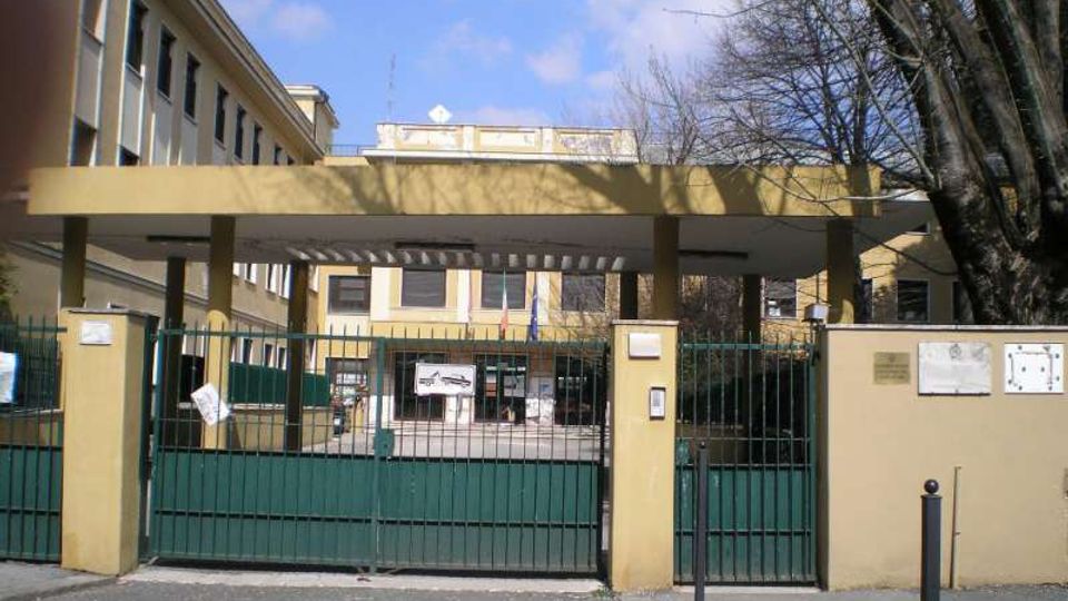 Ingresso di un edificio pubblico con cancello metallico verde e pilastri gialli, coperto da una tettoia orizzontale che conduce al cortile interno. Sullo sfondo si vede la facciata dell’edificio con finestre e bandiere, affiancata da alberi spogli lungo il perimetro.