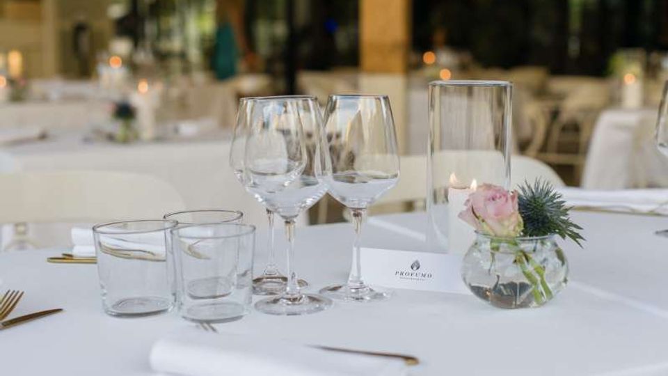 Tavolo apparecchiato in un ristorante elegante con tovaglia bianca, bicchieri da vino e da acqua, posate dorate e un piccolo centrotavola floreale in vaso di vetro. Sullo sfondo si intravedono altri tavoli e grandi vetrate in un ambiente luminoso.