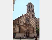 Facciata di una chiesa romanico-gotica in pietra a fasce orizzontali bianche e scure, con grande rosone centrale e portale ad arco. Sul lato si innalza un campanile quadrato a più livelli, affacciato su una piazza urbana con edifici circostanti.