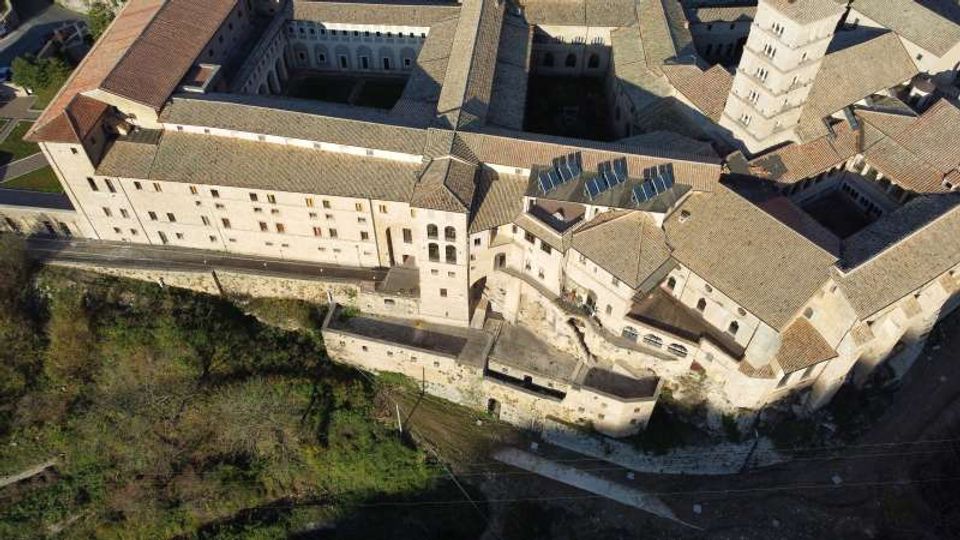 Veduta aerea di un grande complesso storico in pietra con tetti in tegole, cortili interni e un’alta torre campanaria. L’edificio si affaccia su un pendio con mura di contenimento e vegetazione alla base.