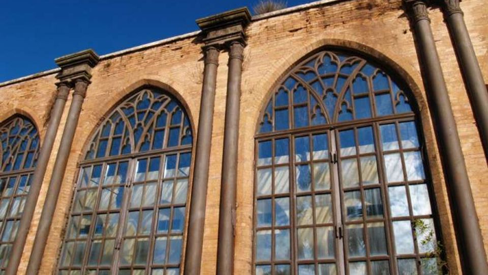 Facciata in mattoni di un edificio storico con alte finestre ad arco a sesto acuto e vetrate a riquadri con motivi gotici nella parte superiore. Pilastri verticali scuri scandiscono la parete sotto un cielo azzurro.