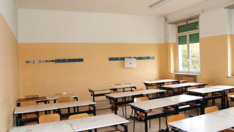 Aula scolastica con banchi e sedie disposti in file, pareti bicolori (bianco e beige) e lampade fluorescenti a soffitto. Sul lato destro ci sono grandi finestre con tapparelle verdi che illuminano la stanza.