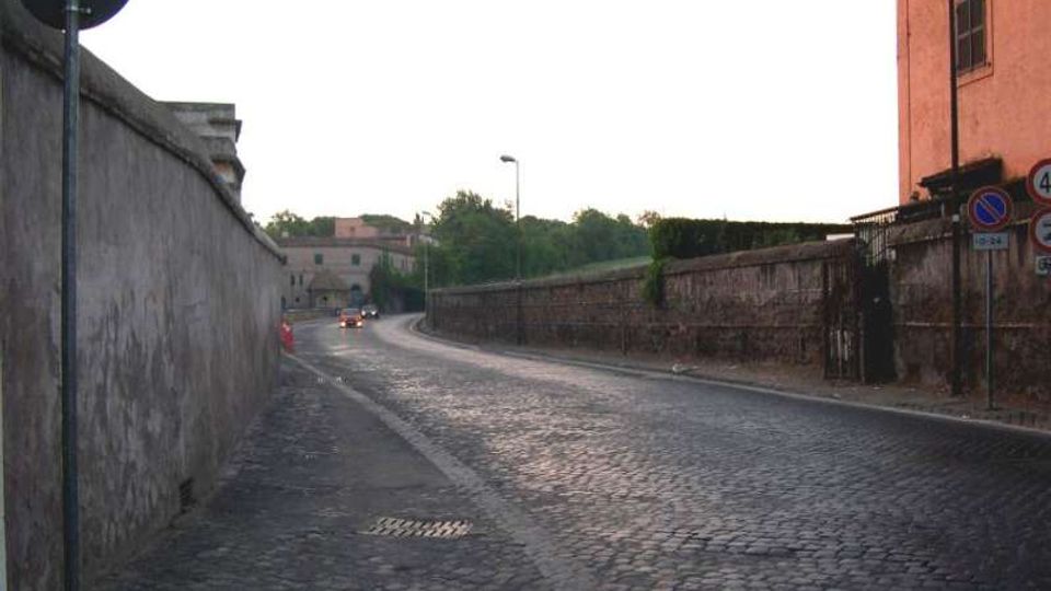 Strada urbana in pavé che curva leggermente, affiancata da alti muri in muratura e intonaco con segnaletica stradale. Sullo sfondo si intravedono edifici e alberi lungo il margine della carreggiata.