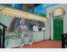 Murale su una parete esterna che raffigura una scena di ristorante con panchina, cameriere e tavolo apparecchiato sotto una tenda dipinta. Accanto c’è una porta verde con insegna “Ristorante Pericchi”, una lampada a muro e pavimentazione in piastrelle di terracotta.