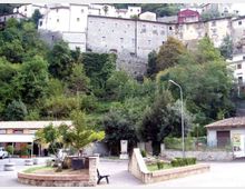Veduta di un borgo collinare con case in pietra e intonaco addossate al pendio, sopra una fitta vegetazione di alberi e cespugli. In primo piano si vede una piccola piazza con aiuole, una panchina, muretti in pietra e un lampione.
