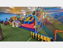 Area giochi interna per bambini con pavimento in erba sintetica, gonfiabili colorati con scivolo e un grande percorso gioco a rete con trampolini. Sullo sfondo si vedono casette e piccoli scivoli, sotto un soffitto con pannelli e luci al neon.