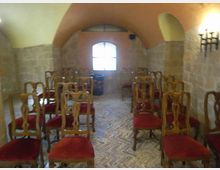 Piccola sala interna con soffitto a volta e pareti in pietra, arredata con file di sedie in legno con seduta rossa su pavimento in mattoni a spina di pesce. In fondo c’è una finestra ad arco che illumina l’ambiente e una lampada a parete in ferro.