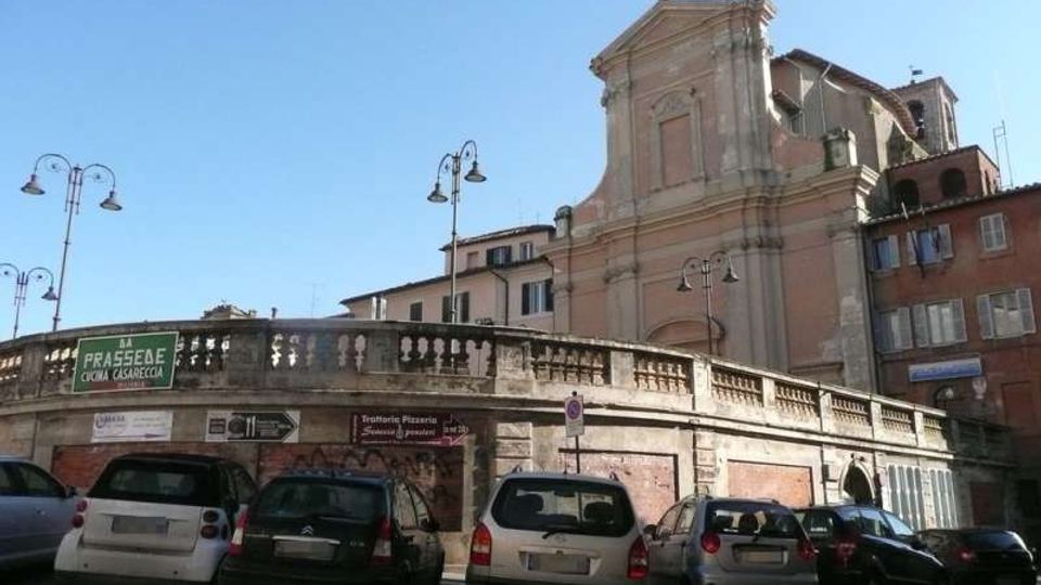 Vista urbana con un ponte in muratura con balaustra in pietra e lampioni, davanti a un parcheggio su strada. Sullo sfondo si vede una chiesa in stile barocco con facciata alta e edifici residenziali adiacenti sotto un cielo sereno.