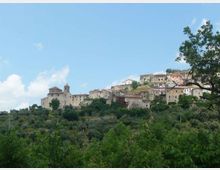 Panorama di un borgo collinare con case in pietra e una chiesa con campanile, disposto a terrazze sul crinale. In primo piano si vede una distesa di vegetazione e alberi, con cielo azzurro sullo sfondo.