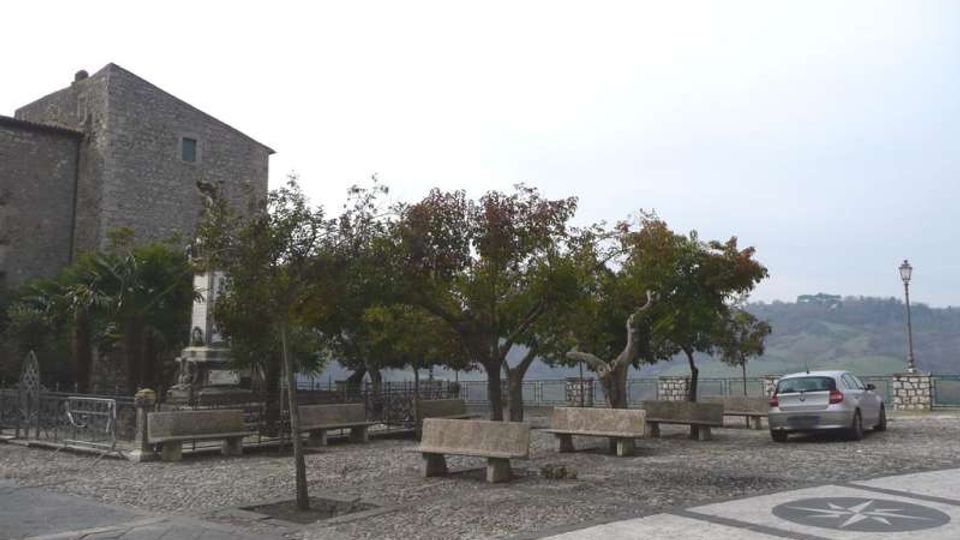 Piazzetta panoramica con pavimentazione in pietra e panchine in cemento sotto alcuni alberi, affacciata su un paesaggio collinare. Sul lato sinistro si vede un edificio storico in muratura e, sullo sfondo, una ringhiera in pietra con un lampione e un’auto parcheggiata.