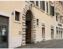 Facciata di edificio storico in una via urbana, con grande portale ad arco in pietra bugnata e porta in legno scuro. Al piano strada si vedono vetrine di negozi e finestre con inferriate, mentre ai piani superiori ci sono finestre con persiane verdi.