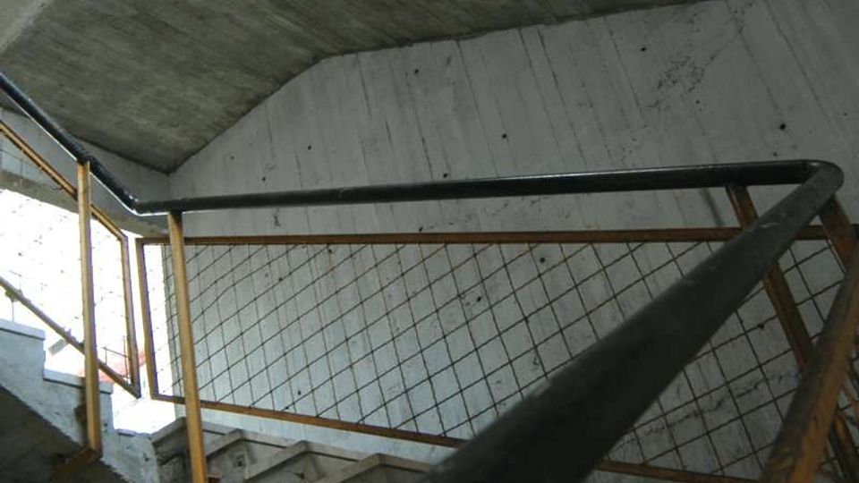 Interno di una tromba delle scale in cemento grezzo, con rampe sovrapposte e pianerottoli angolari. Ringhiere metalliche con rete e corrimano scuro in primo piano lungo il percorso.