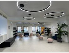 Ampia sala d’attesa moderna con pavimento chiaro, pareti bianche e grandi lampade circolari a LED sospese. Sono presenti poltroncine e divanetti dai colori neutri, scaffalature laterali, piante in vaso e finestre sul fondo che illuminano l’ambiente.