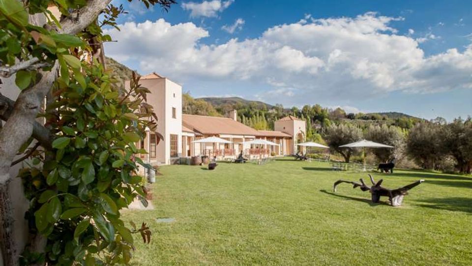 Ampio prato verde davanti a un complesso di edifici chiari con tetti in tegole e portico, in un contesto rurale con colline sullo sfondo. Sul prato ci sono ombrelloni e arredi da esterno, circondati da alberi e vegetazione.